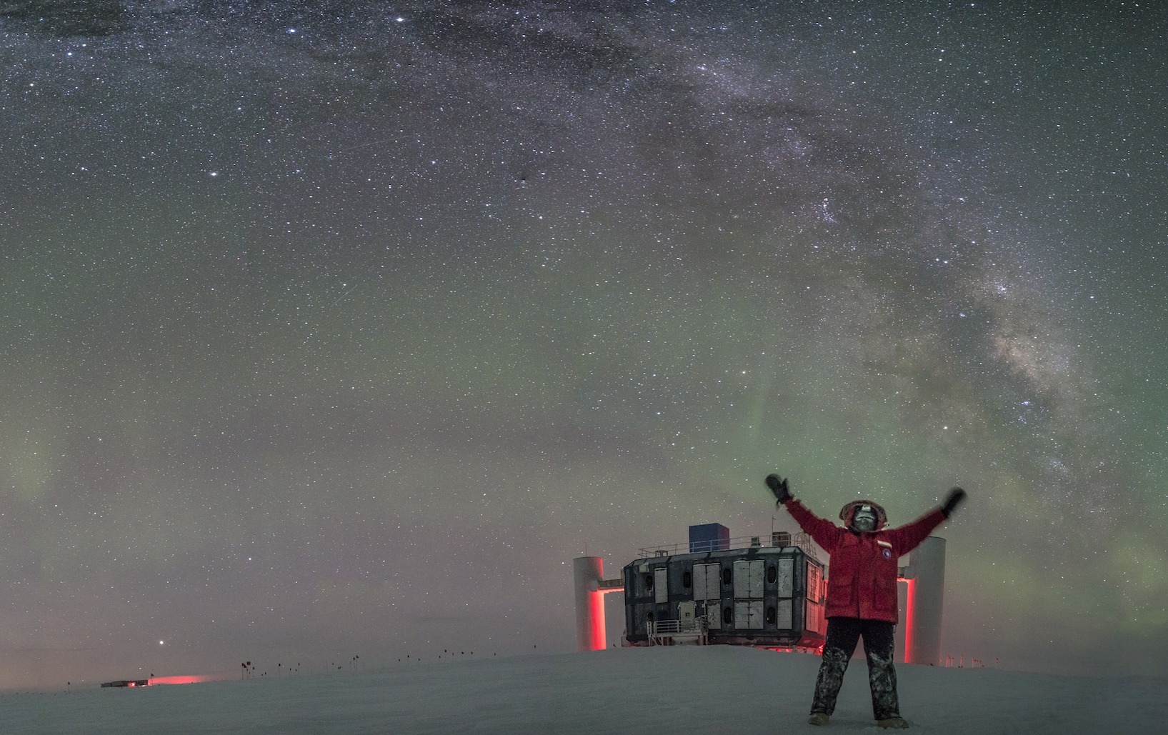 Forscher im roten Polaranzug, im Hintergrund das IceCube-Laborgebäude bei Nacht vor Sternenhimmel