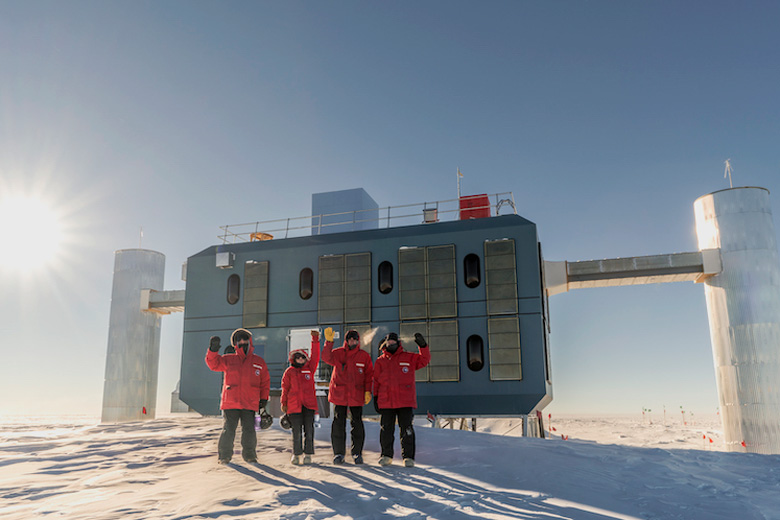 Vier Personen in roten Polaranzügen vor dem IceCube-Laborgebäude am Südpol, im Hintergrund strahlend blauer Himmel